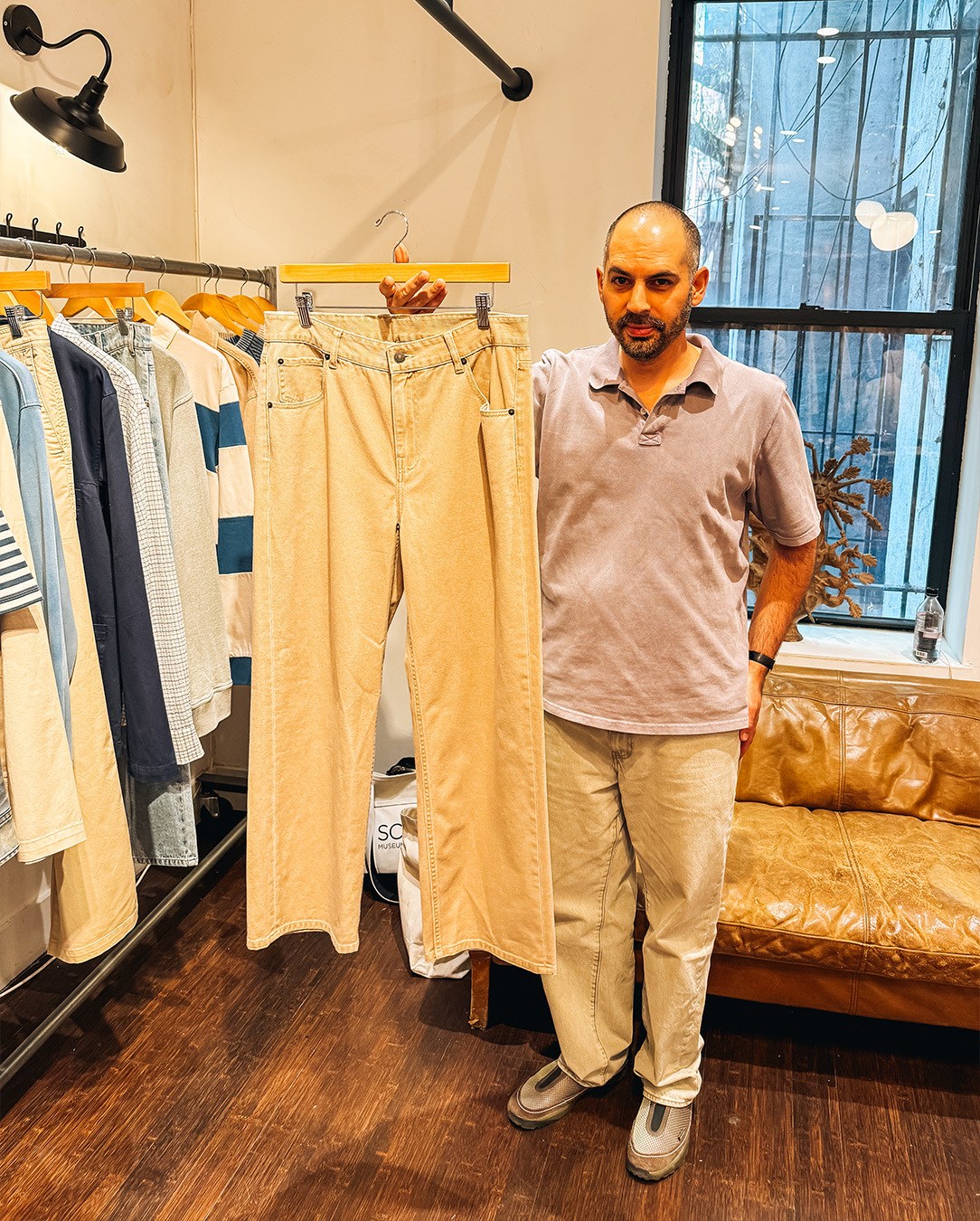 Man in a purple polo and tan pants holds up a pair of tan wide-leg jeans in a clothing store.