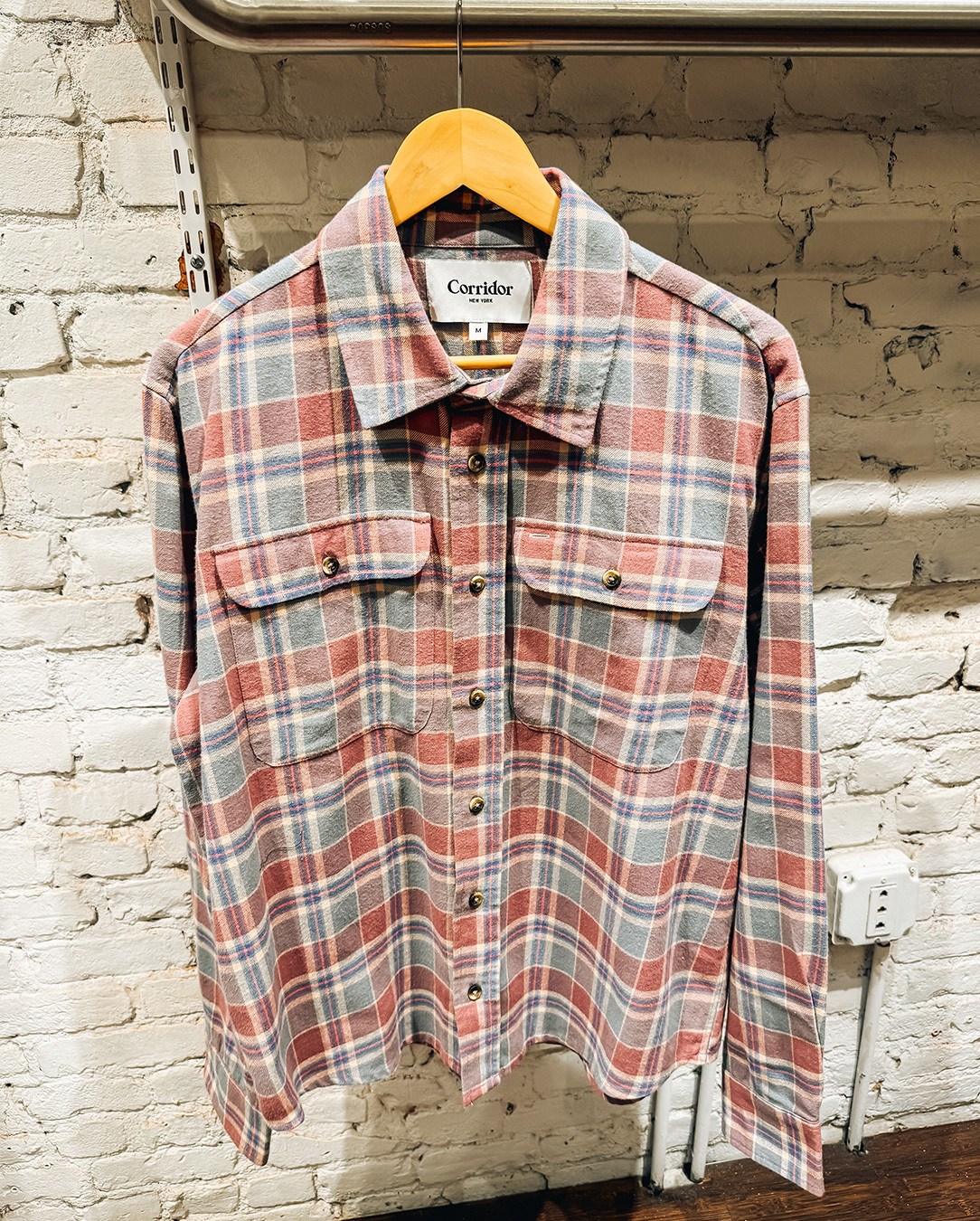 Corridor New York flannel shirt in red, blue, and cream plaid, hanging on a wooden hanger against a white brick wall.