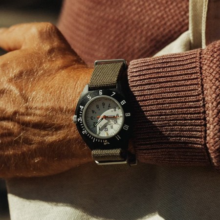 A person's arm with a black watch, white face, and olive green strap, wearing a rust-colored sweater.
