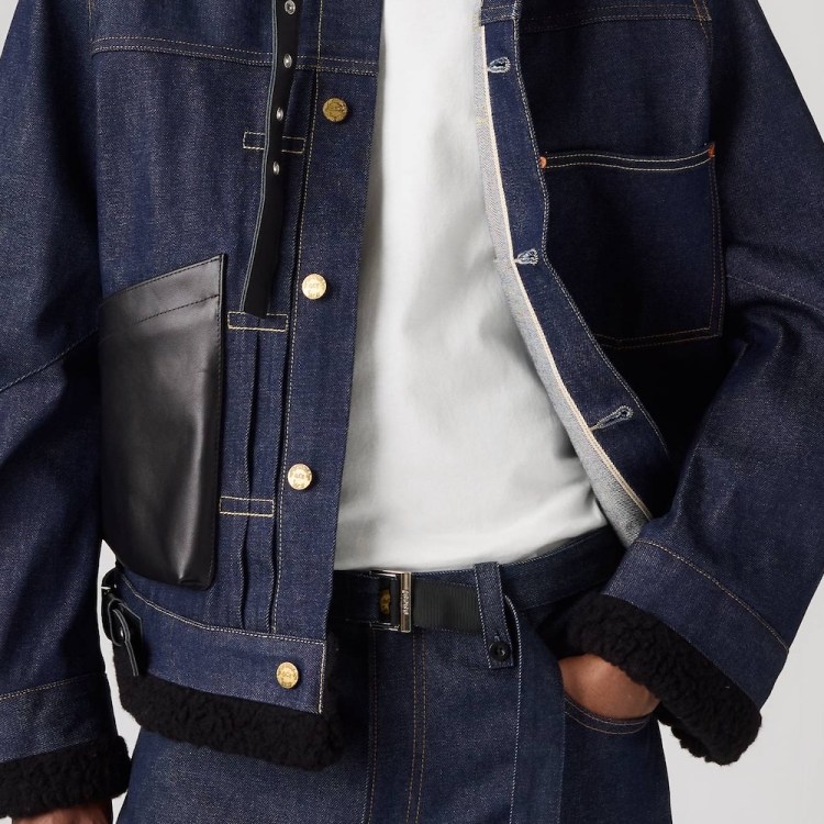 Person wearing a dark denim jacket with gold buttons, a black leather pocket, and black fleece trim, over a white t-shirt.