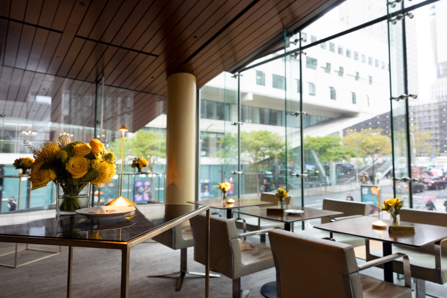 Modern restaurant interior with floor-to-ceiling windows overlooking a city street, tables set with yellow flowers.
