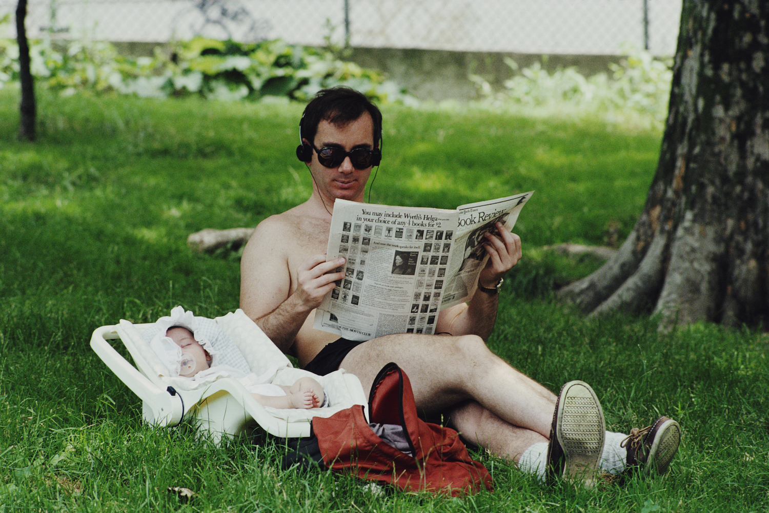 A man in sunglasses and headphones reads a newspaper in the grass next to a sleeping baby in a car seat.