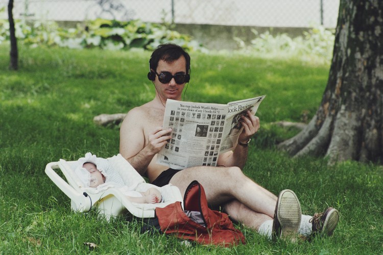 A man in sunglasses and headphones reads a newspaper in the grass next to a sleeping baby in a car seat.