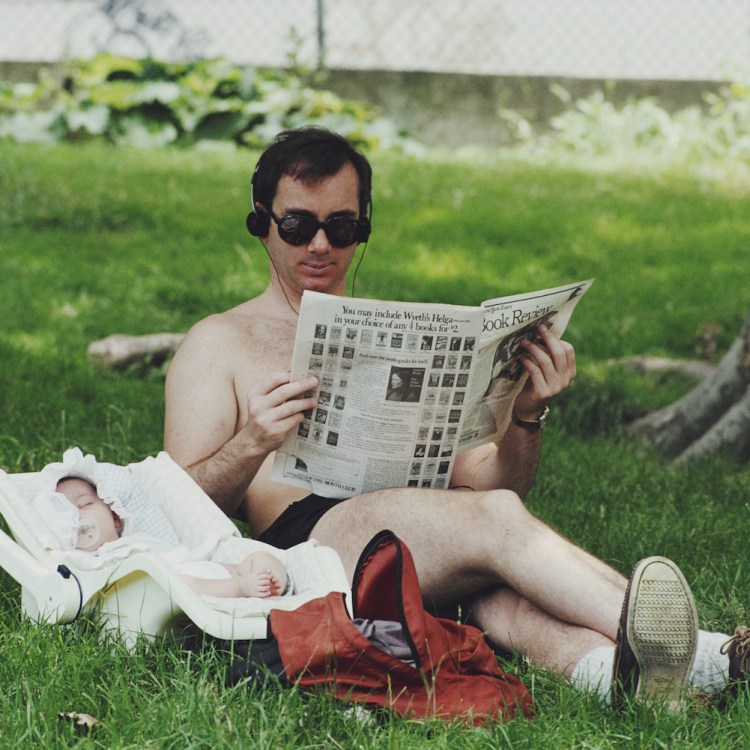 A man in sunglasses and headphones reads a newspaper in the grass next to a sleeping baby in a car seat.