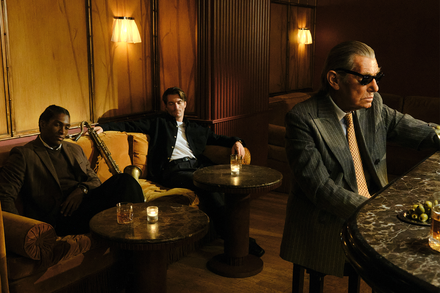 Three men in a dimly lit bar: two seated on a couch with a saxophone, one in sunglasses at a marble bar.
