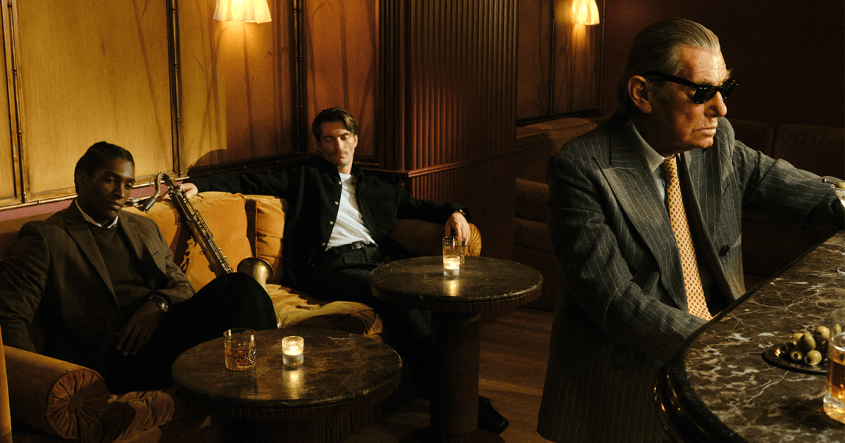 Three men in a dimly lit bar: two seated on a couch with a saxophone, one in sunglasses at a marble bar.