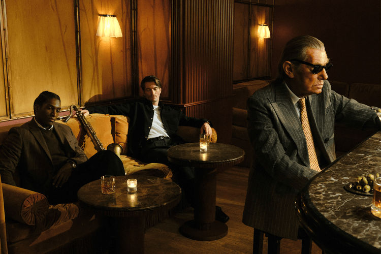 Three men in a dimly lit bar: two seated on a couch with a saxophone, one in sunglasses at a marble bar.