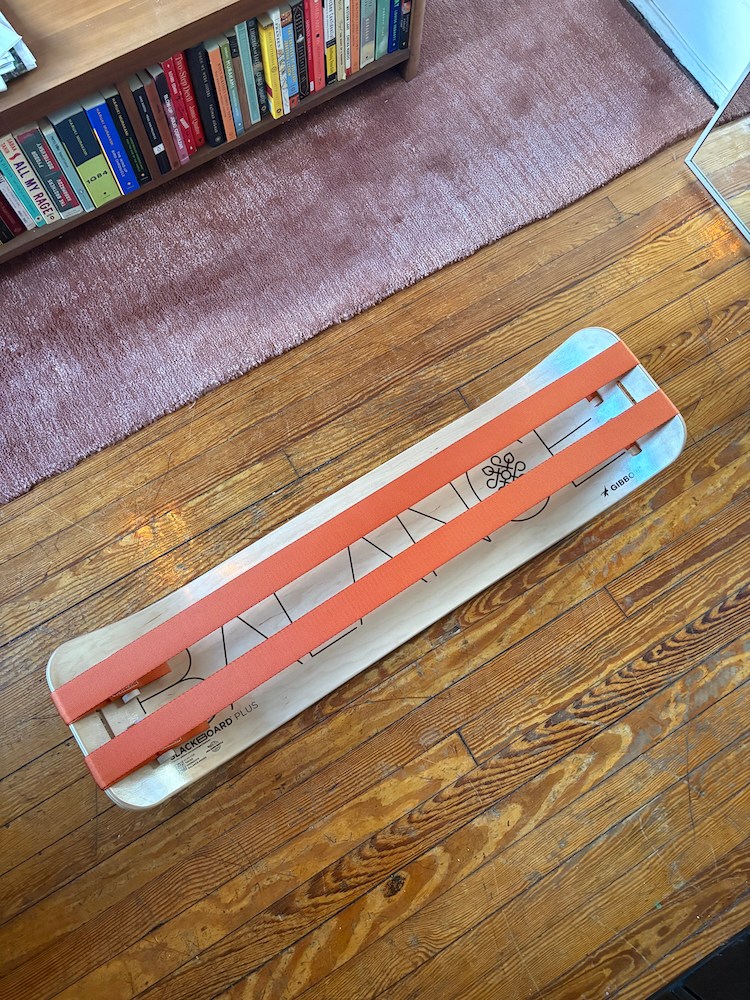 A wooden balance board with two orange straps on a hardwood floor, with a pink rug and bookshelf in the background.
