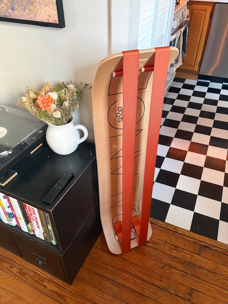 A wooden balance board with orange straps leans against a wall next to a record player and dried flowers.